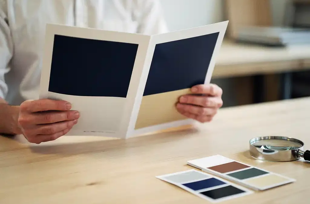Kwaliteitscontroleur vergelijkt twee gedrukte brochures in Amsterdam studio met kleurstalen en loepvergrooter op lichte eiken tafel.