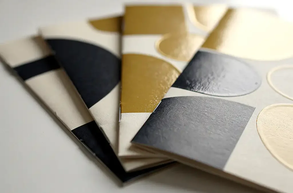 Marketingbrochures uitgewaaierd op wit bureau met glanzende, matte en geëmbosseerde afwerkingen in crème, antraciet en goud.